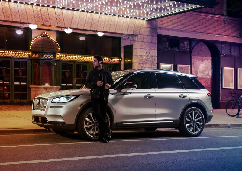 A 2022 Lincoln Corsair SUV is parked outside a theater as the driver relaxes against the frame and lights illuminate the floating roofline and body | Performance Lincoln Bountiful in Bountiful UT