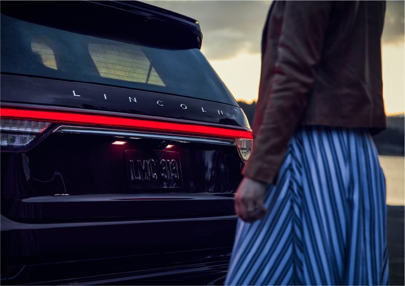 A person is shown near the rear of a 2023 Lincoln Aviator® SUV as the Lincoln Embrace illuminates the rear lights | Performance Lincoln Bountiful in Bountiful UT